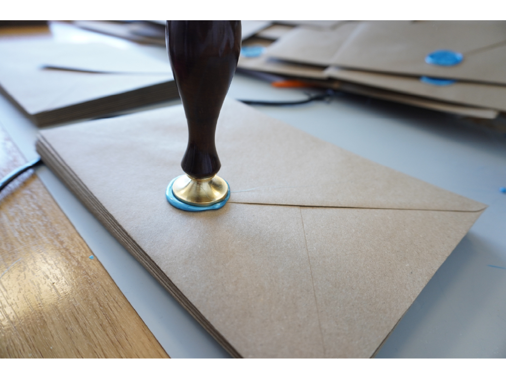 A kraft invitation envelope being hand-sealed with blue wax and a brass stamp